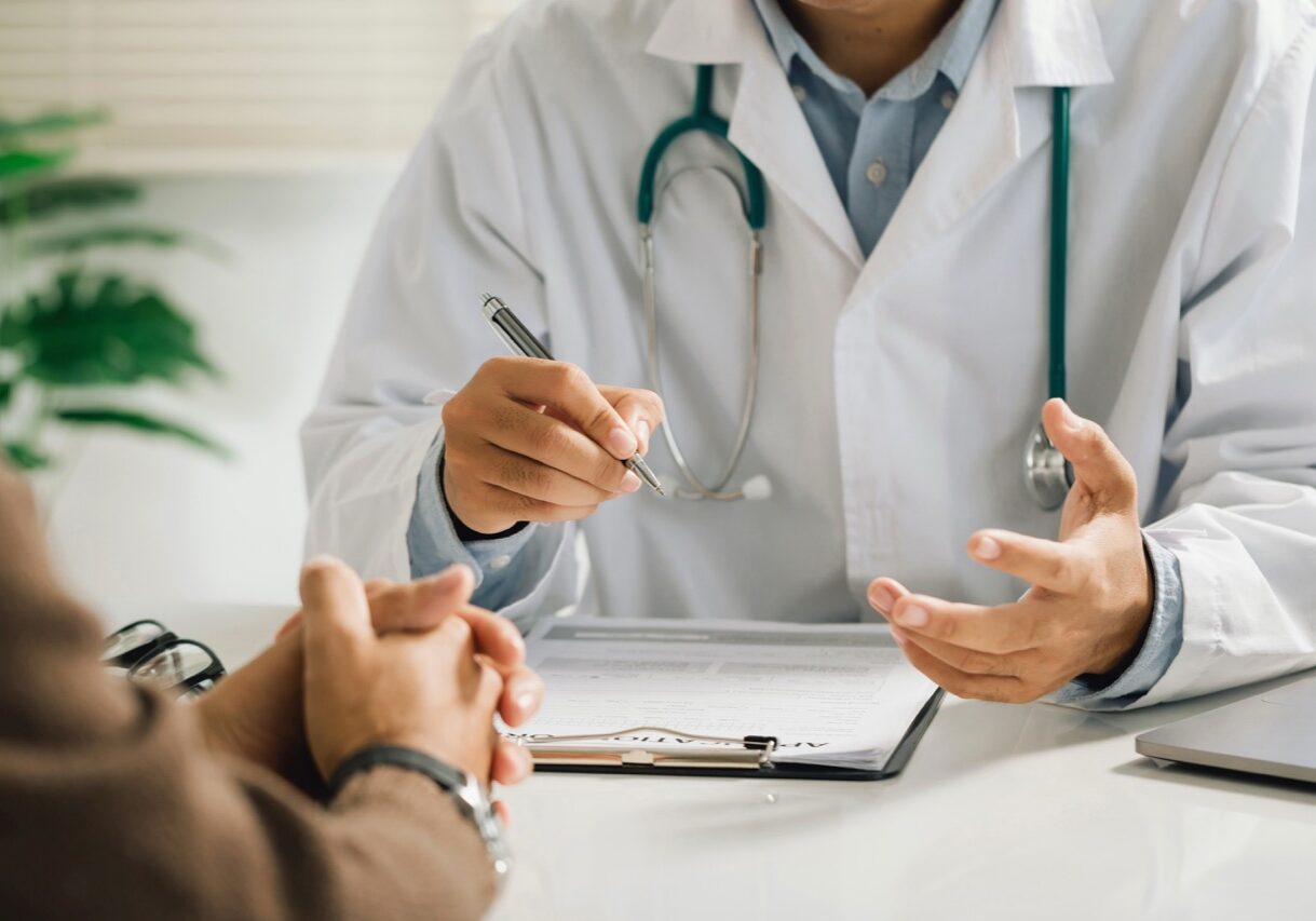 A professional physician in a white medical uniform talks to discuss results or symptoms and gives a recommendation to a male patient and signs a medical paper at an appointment visit in the clinic.